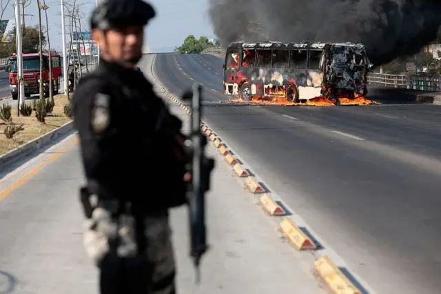 Un autobús incendiado al fondo y un policía en primer plano, en una calle de la zona metropolitana de Guadalajara