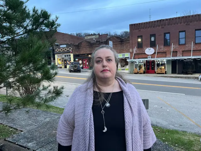 Una mujer con el pelo gris y una rebeca morada está de pie frente a una hilera de tiendas en la plaza principal de Gainesboro.