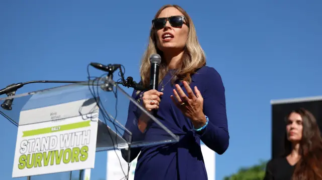 Annie Farmer, con un micrófono en la mano, habla en un atril. Lleva gafas de sol y viste de azul. Hace sol y el atril muestra el mensaje "Junto a las víctimas" escrito en inglés.