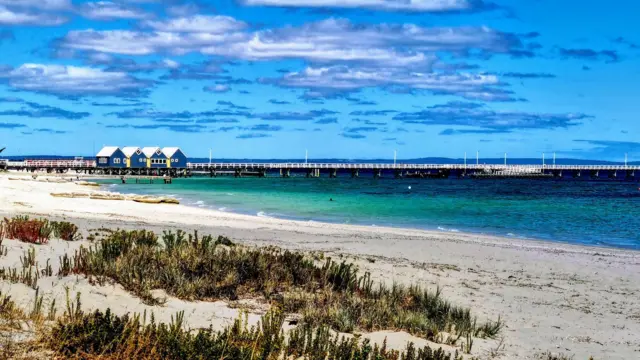 Se ve una playa de un azul brillante con cuatro cabañas a lo largo de un muelle. Es un día soleado.