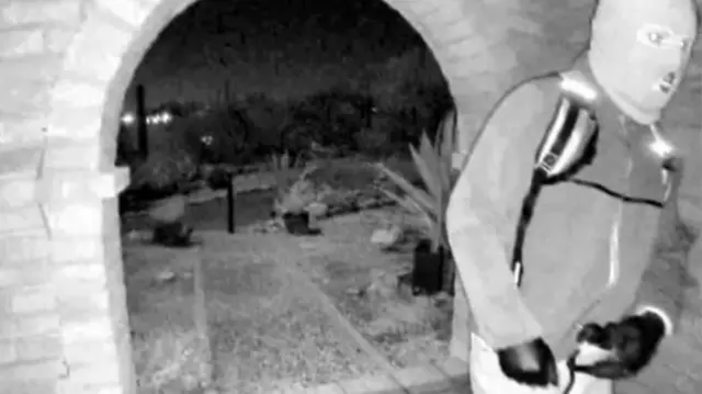 La policía ha publicado imágenes en blanco y negro de una persona enmascarada, en las que se ve a la persona con guantes y un jersey con cremallera acercándose a la casa por la noche.