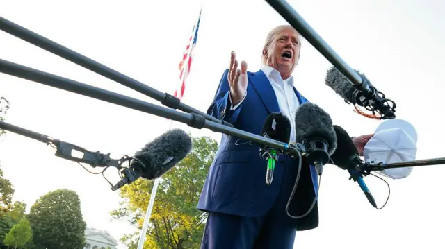 Rodeado de micrófonos y con una bandera estadounidense en un asta detrás de él, el presidente Donald Trump habla con periodistas afuera de los edificios de la Casa Blanca, vistiendo un traje azul y una camisa blanca de cuello abierto y sosteniendo una gorra blanca en su mano izquierda.