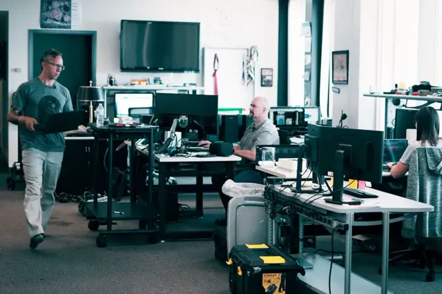 Greg Squire y sus colegas, entre ellos Pete Manning, en su oficina. Greg lleva una camiseta verde y pantalones claros y cruza una sala llena de pantallas de computadora con una computadora portátil en la mano. 