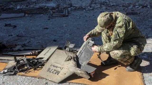 Un agente de policía vestido con uniforme militar se agacha para inspeccionar los restos de lo que se cree que es un dron ruso del tipo Shahed. 