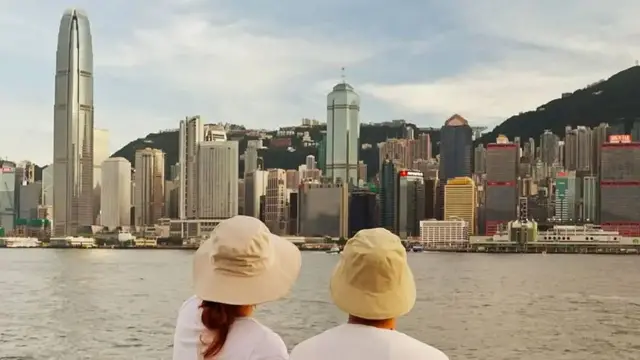 Eric y Emily de espaldas con sombreros sentados junto a un río. Al otro lado del río se ven edificios y rascacielos.
