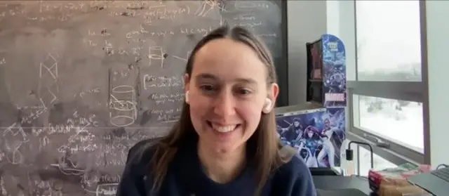 Sabrina González Pasterski, con una camisa azul oscura, sonriendo. Detrás tiene un pizarrón lleno de fórmulas