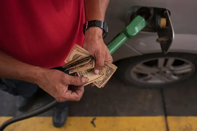 Un hombre cuenta dólares en una estación de gasolina. La imagen muestra sus manos, los dólares y al fondo se ve un auto con un surtidor de gasolina.