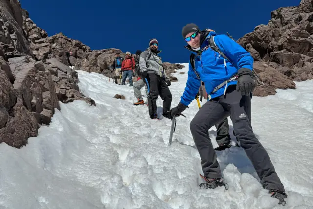 La foto muestra la ladera de una montaña nevada, con algunas rocas escarpadas a la vista y un cielo azul profundo y despejado. La foto se tomó desde abajo, con Gerwyn de pie en primer plano y otros cinco hombres visibles en distintos grados al fondo. Gerwyn se apoya en un piolet clavado en la nieve. Todos llevan ropa de abrigo y gafas de sol.