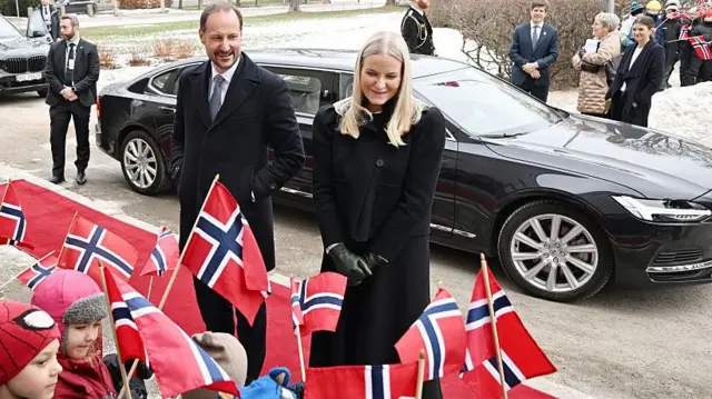 Una pareja vestida con abrigos negros está de pie delante de un coche, mientras unos niños en primer plano agitan banderas de Noruega.