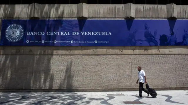 Un hombre con una camisa blanca camina por la acera frente a una pared con un letrero que dice "Banco Central de Venezuela".