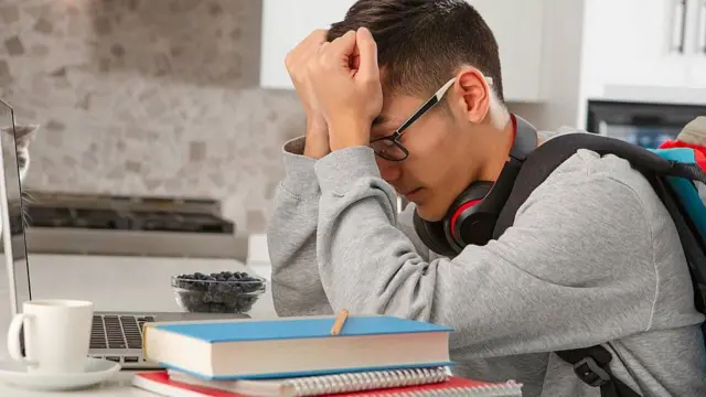 Joven con los ojos cerrados se agarra la cabeza con las manos en señal de frustración mientras está sentado frente a libros, cuadernos y una computadora