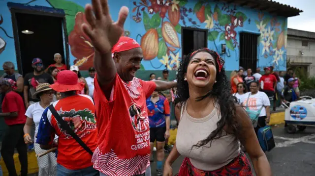 Una mujer muy sonriente baila, a su lado, un hombre vestido con una camiseta roja, baila también tambor.