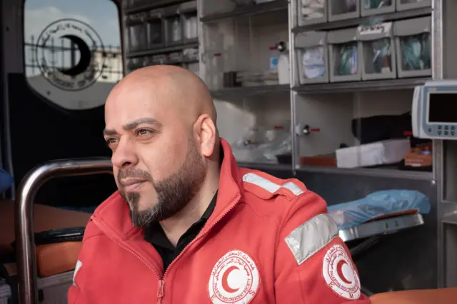 El paramédico Hassan Fouqha está sentado en el borde de la puerta abierta de la ambulancia, con una chaqueta roja de médico, mirando al frente.