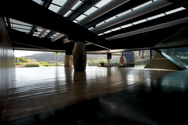 Interior sombreado con techo estructural visible, grandes vigas y columnas de piedra, abierto hacia un paisaje exterior iluminado.