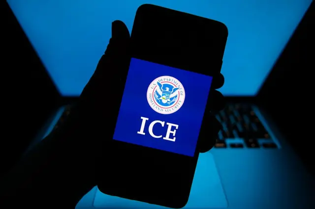 Computadora en el fondo y en primer plano una mano sosteniendo un teléfono con el logo de ICE en la pantalla
