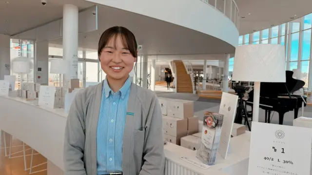 Riona Okada, en un gran salón decorado en tonos blancos, sonríe a la cámara. Viste una blusa azul claro y una chaqueta gris.