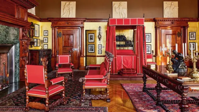 El dormitorio de George W. Vanderbilt, con una gran mesa con candelabros, sillones rojos en torno a una estufa a leña y frisos con esculturas griegas en la parte superior de las paredes.