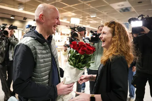 Un hombre sonriente recibe a una mujer sonriente con un ramo de rosas, mientras camarógrafos graban el momento.