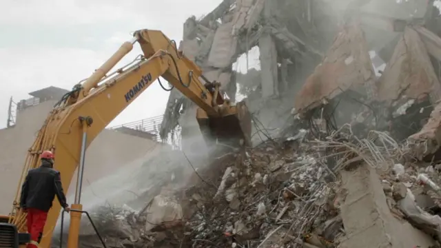 Los restos de un edificio, compuestos por hormigón destrozado y metal retorcido. Un hombre, vestido con chaqueta negra, pantalones naranjas y casco de seguridad, maneja una excavadora para remover los escombros.