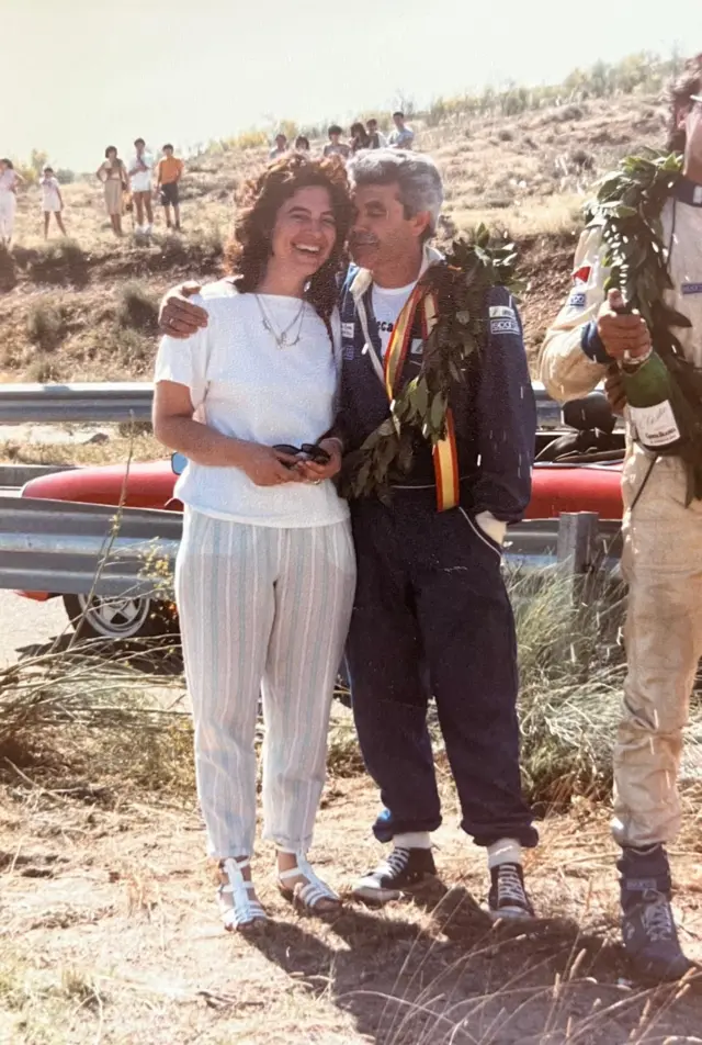 Juan, victorioso con una medalla al cuello y una corona de laureles, abraza con cariño a su esposa quien sonríe alegremente a la cámara.