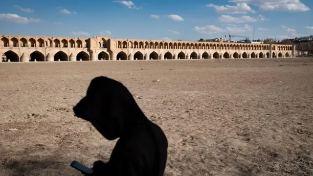 Un hombre iraní utiliza su teléfono móvil mientras permanece de pie en la orilla seca del río Zayandeh Rud, con el histórico puente Si-o-se-pol (Puente de los 33 Arcos) visible al fondo, en la histórica ciudad de Isfahán, Irán, a principios de 2025.