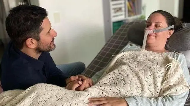 Paola Roldán, incorporada en una cama, con un respirador en la nariz, tapada con una manta y agarrada de la mano de su esposo, a la izquierda y de perfil. 
