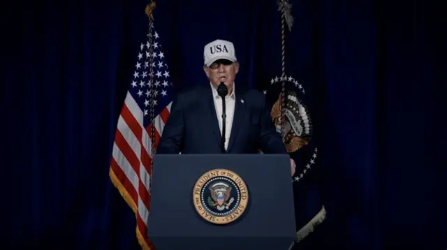 Una captura de pantalla de un video publicado en la cuenta de Truth Social del presidente estadounidense Donald Trump lo muestra pronunciando unas declaraciones sobre las operaciones de combate contra Irán el 28 de febrero de 2026. Lleva un traje azul marino y una gorra blanca con el logotipo de EE. UU., con la bandera estadounidense y la bandera presidencial visibles al fondo.