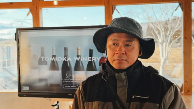 Junichiro Hosokawa, gerente da Tomioka Winery, con gesto serio, posa delante de una imagen con botellas de vino