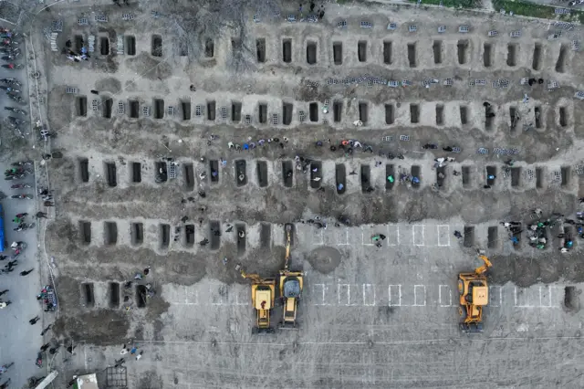 Una fotografía aérea muestra una serie de tumbas excavadas en el suelo con personas alrededor. También se ven tractores amarillos.