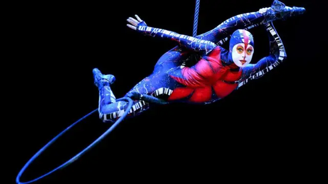 Una artista del Cirque du Soleil, con un traje azul y rojo y maquillaje completo, se balancea en un trapecio mientras actúa con un aro de hula hula.