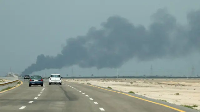 Tres coches avanzan por una autopista de tres carriles con humo frente a ellos. El cielo tiene un tono azul oscuro y los laterales de la carretera muestran polvo y algunos arbustos dispersos.