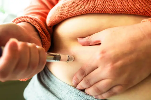 Primer plano de las manos de una mujer sosteniendo una pluma de inyección para la pérdida de peso. Ella se está inyectando la dosis en el abdomen.