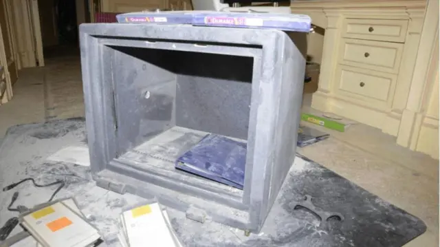 Una caja fuerte con la puerta rota sobre un suelo alfombrado, con polvo esparcido por todas partes y lo que parecen ser muebles de dormitorio de color crema con cajones incorporados en el fondo.