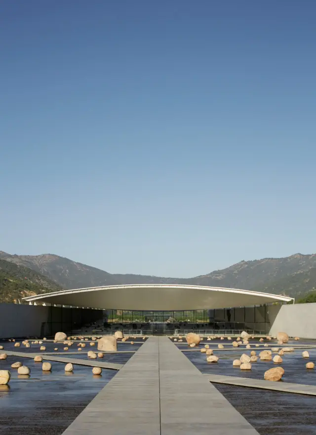 Pasarela de hormigón que conduce a un edificio de techo curvo, rodeado de un estanque con numerosas rocas y montañas al fondo bajo un cielo despejado.