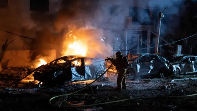 Un bombero dirige agua con una manguera hacia un coche en llamas durante la noche; el vehículo arde con un intenso tono naranja y otros coches a su alrededor muestran daños graves.