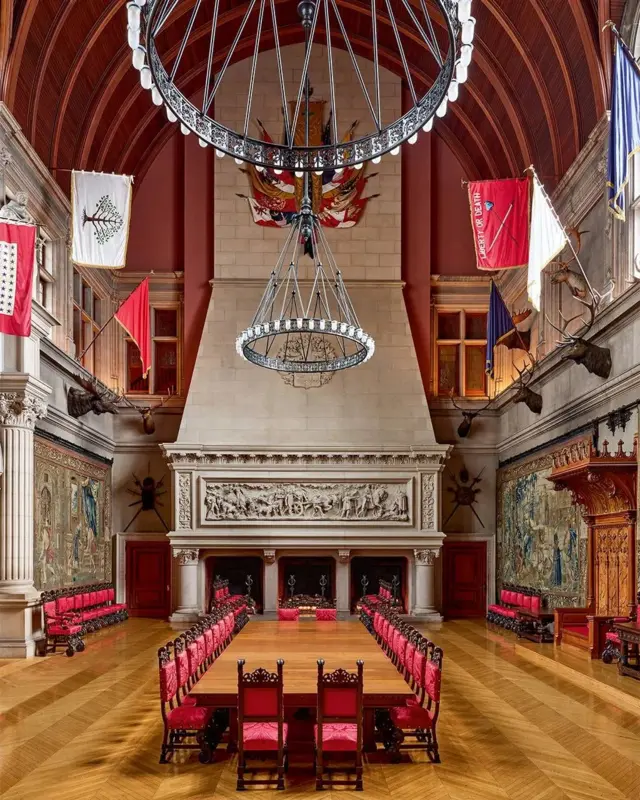 Sala de techos altos y tapices en las paredes. En el centro se ve una larga mesa de madera para banquetes con sillas tapizadas en rojo para cerca de 20 comensales.