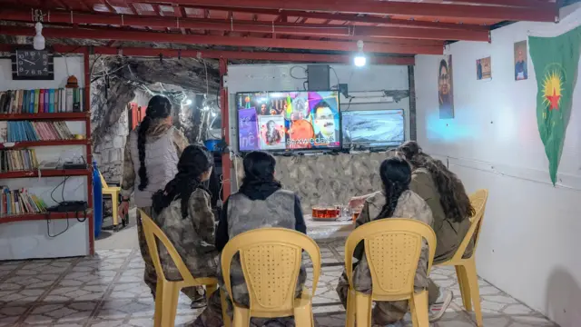 Cinco combatientes sentadas en sillas de plástico viendo la televisión en una habitación improvisada en una cueva.