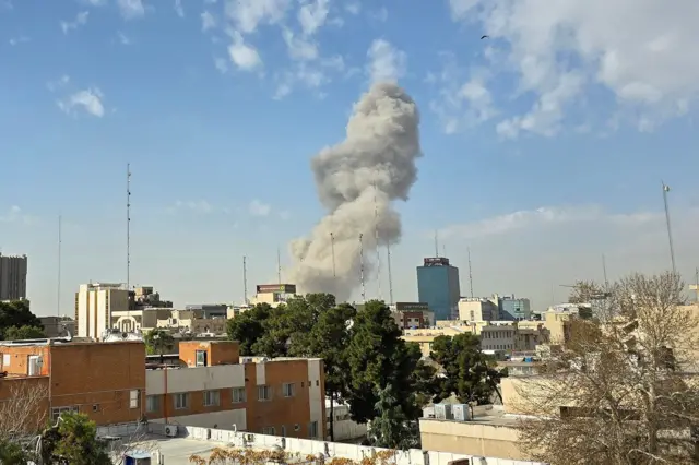Humo sale al fondo de edificios, árboles y lo que parecen ser viviendas