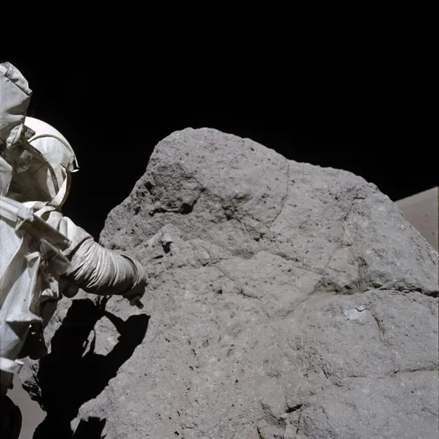 El astronauta Harrison Schmitt, de la misión Apolo 17, está recogiendo muestras de roca en la Luna. La roca es ligeramente más alta que él y aproximadamente tres veces más ancha. Aparece a la izquierda de la foto, de espaldas a la cámara, y lleva un traje espacial blanco de la NASA mientras intenta recoger la muestra con el brazo derecho. Su cuerpo proyecta una sombra sobre la gran roca de color gris oscuro. En la parte superior de la foto se puede ver la oscuridad del espacio.