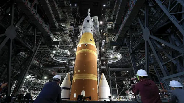 El Artemis II se encuentra en el Edificio de Ensamblaje de Vehículos del Centro Espacial Kennedy de la NASA, mientras tres trabajadores con cascos blancos miran hacia el cohete