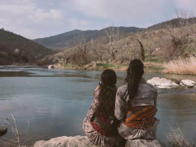 Delal (izquierda) descansa con una compañera combatiente cerca de un pequeño lago cerca de su base. Están de espaldas a la cámara y visten la vestimenta tradicional kurda.