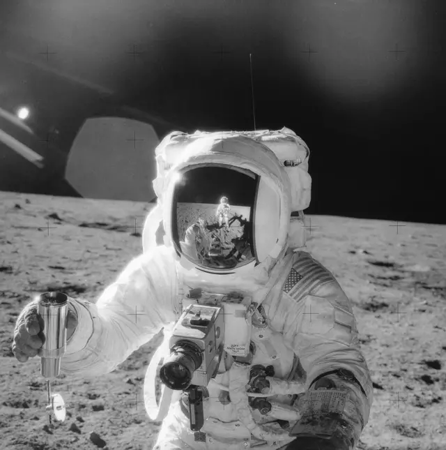 Fotografía en blanco y negro de la misión Apolo 12 de 1969 en la que se ve a uno de los astronautas en la superficie lunar sosteniendo un recipiente con suelo lunar. En el casco se refleja la imagen del otro astronauta.