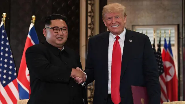 A la izquierda, Kim Jong-un, ataviado con un traje oscuro de estilo "Mao"; a la derecha, Trump, vistiendo un traje negro y corbata roja. Ambos líderes aparecen sonriendo y estrechándose la mano frente a las banderas de Estados Unidos y Corea del Norte.
