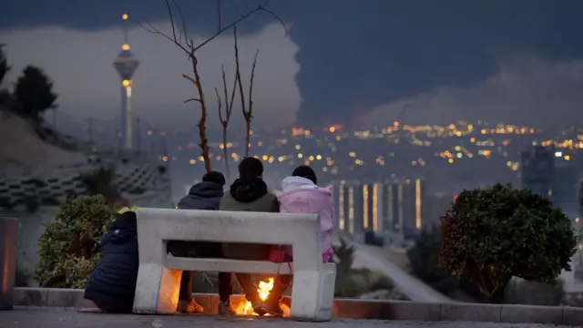 Tres personas en Teherán observan cómo arden los incendios en el depósito de petróleo de Shahran el 8 de marzo, mientras ataques israelíes tenían como objetivo centros de combustible cerca de Teherán.