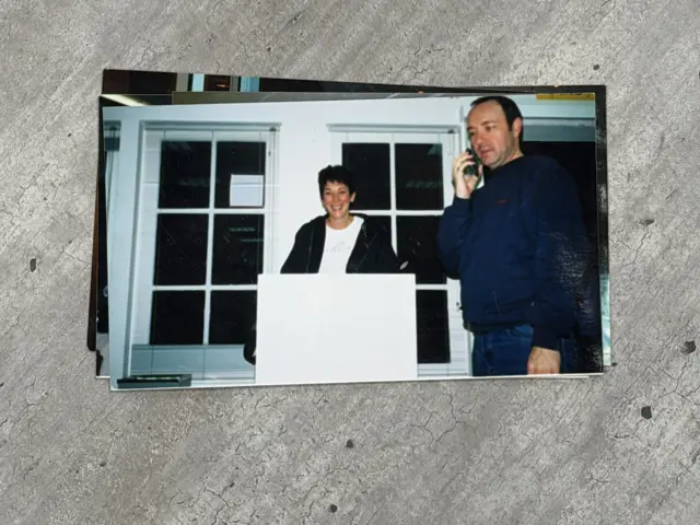 Una mujer, Ghislaine Maxwell, con el pelo corto, sonríe y está de pie detrás de un podio blanco. A su izquierda, un hombre, Kevin Spacey, lleva un teléfono móvil en la oreja derecha. Viste una camisa azul marino y vaqueros azules.