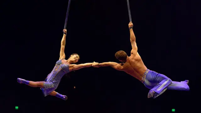 Dos artistas del Cirque du Soleil, una mujer y un hombre con trajes morados iguales, se toman de la mano mientras se balancean en un trapecio durante un espectáculo en agosto de 2025 en Perth, Australia.