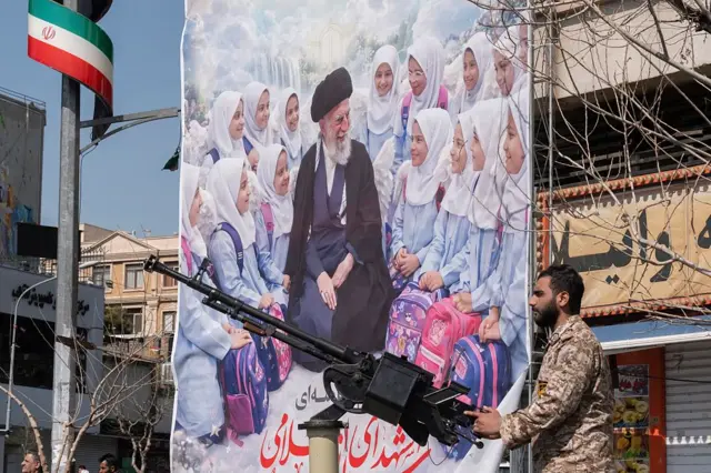 Un miembro militar armado del Cuerpo de la Guardia Revolucionaria Islámica hace guardia detrás de un arma frente a una pancarta que muestra un retrato del difunto líder supremo de Irán, el ayatolá Alí Jamenei, y colegialas.