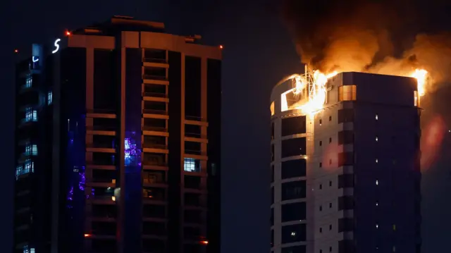 Las llamas envuelven la parte superior de un bloque de viviendas durante la noche mientras el humo se eleva hacia el cielo oscuro. Otro edificio al lado no muestra daños aparentes.