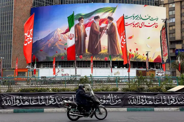 Un hombre pasa en moto junto a una pancarta colgada en la plaza Valiasr, en el centro de Teherán, el 10 de marzo de 2026, en la que aparece el difunto líder supremo de Irán, el ayatolá Ruhollah Jomeini (izquierda), mientras observa cómo su sucesor, el difunto ayatolá Alí Jamenei (en el centro), entrega la bandera nacional a su hijo y nuevo líder supremo, Mojtaba Jamenei.