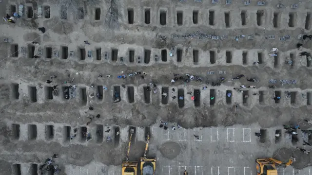 Una foto aérea muestra al menos cinco hileras de agujeros rectangulares en el suelo, algunos señalizados y otros aún sin excavar. Se ven tres excavadoras.